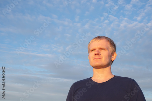 A young Caucasian male blond with a pensive look looks into the distance against a blue sky with light clouds. Sunset light. Copyspace.