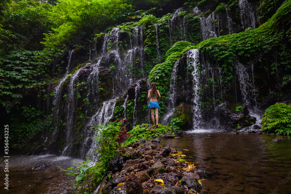 Young traveler woman at waterfall in tropical forest. View from back. Travel concept. Banyu Wana Amertha waterfall Wanagiri, Bali. Slow shutter speed, motion photography.