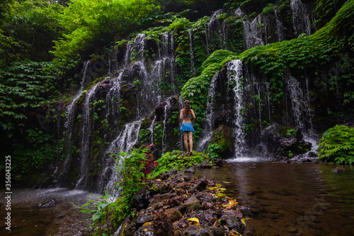 Young traveler woman at waterfall in tropical forest. View from back. Travel concept. Banyu Wana Amertha waterfall Wanagiri, Bali. Slow shutter speed, motion photography.