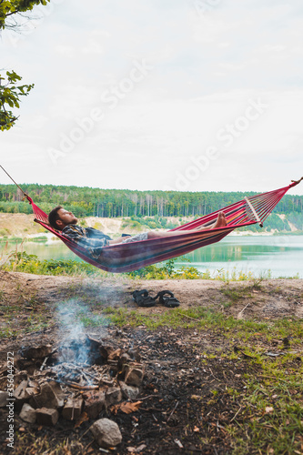 man laying on hammock at lake beach near camp fire