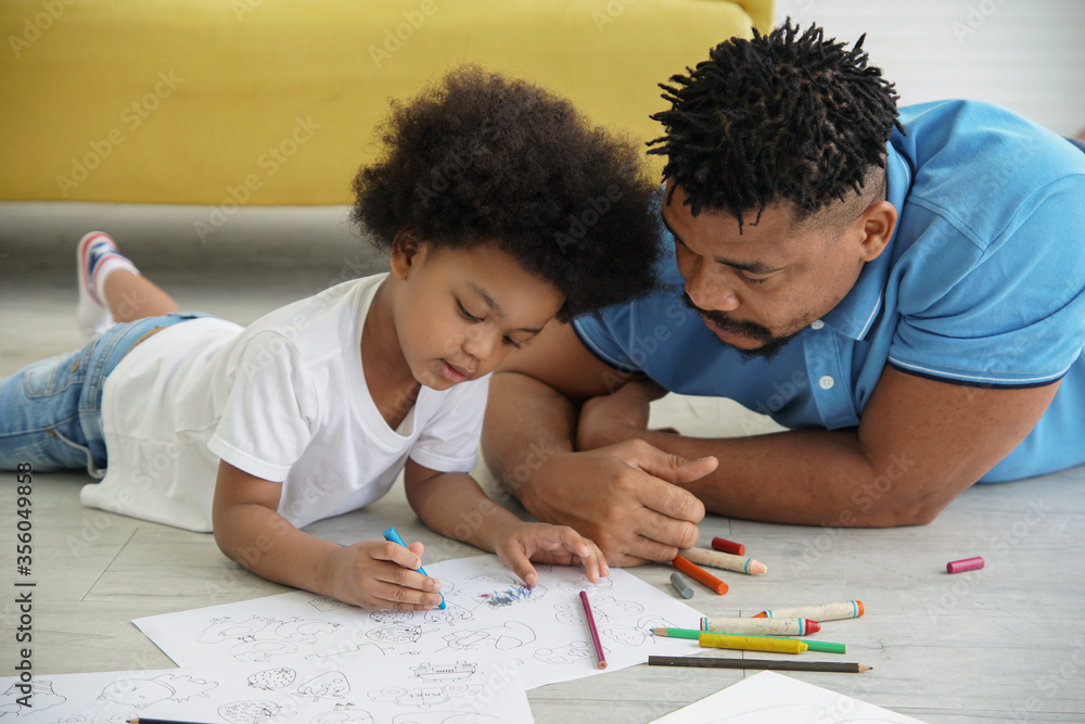 Black African family dad son together at home. Young father and little boy lying on warm floor and drawing coloring on book with color pencil crayon at living room Black African family dad son together at home. Young father and little boy lying on warm floor and drawing coloring on book with color pencil crayon at living room