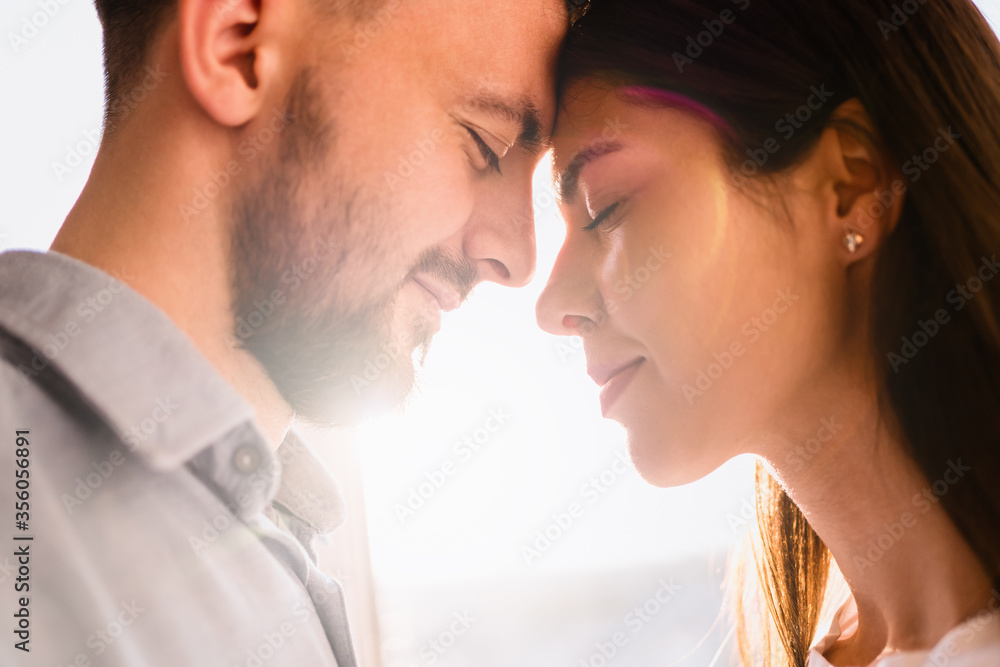 Portrait loving couples enjoying each other company with window in background with sunlight reflection on their faces Portrait loving couples enjoying each other company with window in background with sunlight reflection on their faces