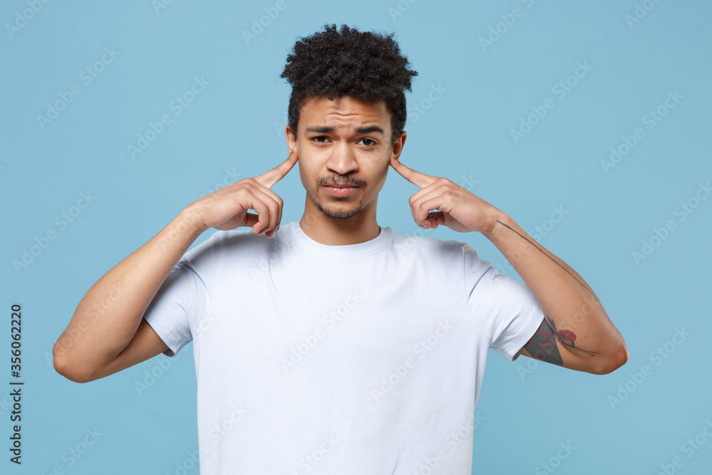 Dissatisfied young african american guy in casual white t-shirt posing isolated on blue background studio portrait. People emotions lifestyle concept. Mock up copy space. Covering ears with fingers. Dissatisfied young african american guy in casual white t-shirt posing isolated on blue background studio portrait. People emotions lifestyle concept. Mock up copy space. Covering ears with fingers.