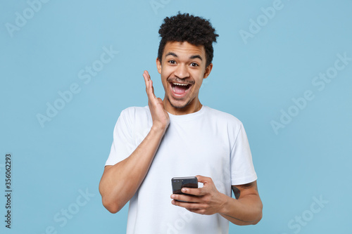Excited young african american guy in casual white t-shirt posing isolated on blue wall background studio portrait. People lifestyle concept. Mock up copy space. Using mobile phone spreading hands.