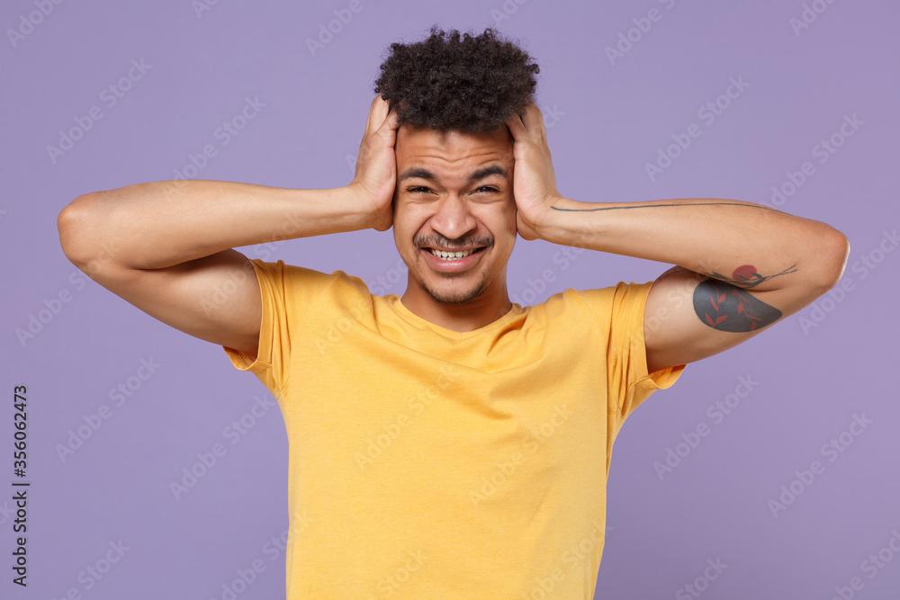 Displeased tired young african american guy in casual yellow t-shirt posing isolated on pastel violet wall background. People lifestyle concept. Mock up copy space. Put hands on head, having headache. Displeased tired young african american guy in casual yellow t-shirt posing isolated on pastel violet wall background. People lifestyle concept. Mock up copy space. Put hands on head, having headache.