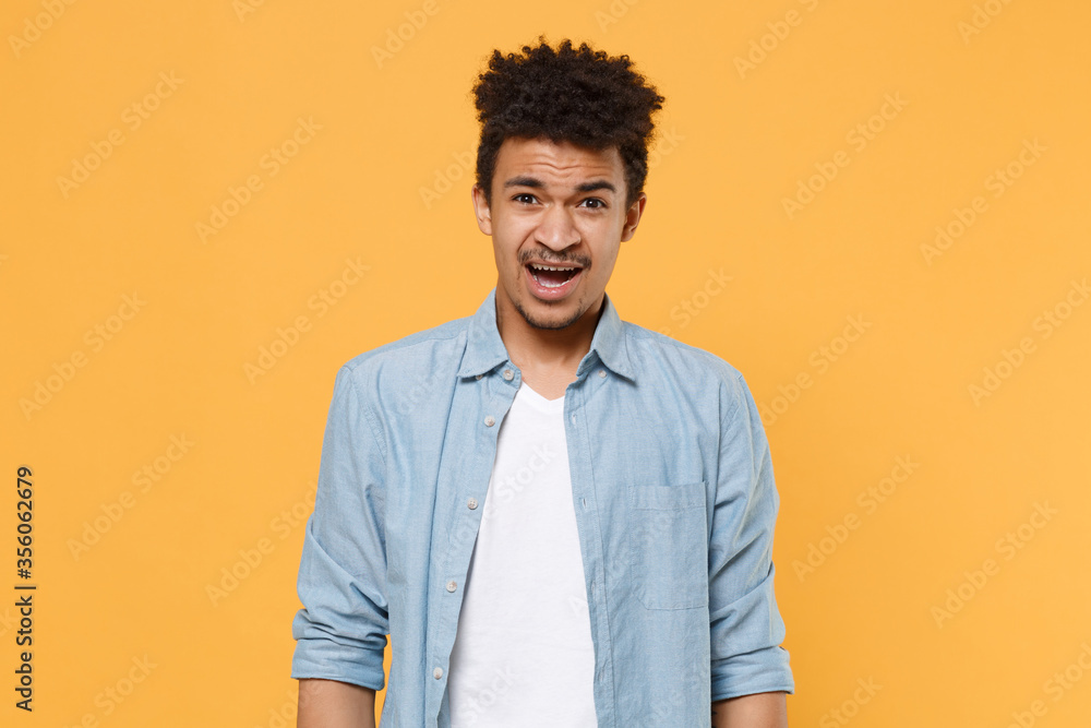 Shocked perplexed young african american guy in casual blue shirt posing isolated on yellow wall background studio portrait. People emotions lifestyle concept. Mock up copy space. Keeping mouth open. Shocked perplexed young african american guy in casual blue shirt posing isolated on yellow wall background studio portrait. People emotions lifestyle concept. Mock up copy space. Keeping mouth open.