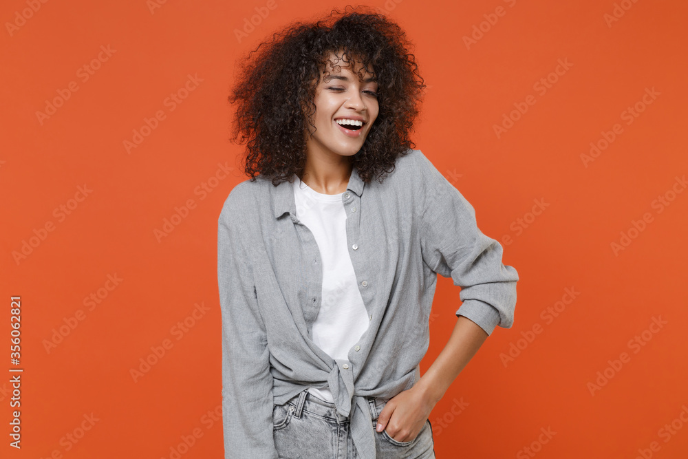 Fototapeta premium Cheerful young african american woman girl in gray casual clothes posing isolated on orange background studio portrait. People lifestyle concept. Mock up copy space. Holding hand in pocket, blinking.
