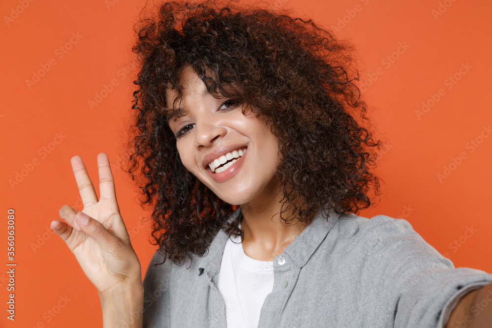 Close up of smiling african american woman in gray casual clothes isolated on orange background. People lifestyle concept. Mock up copy space. Doing selfie shot on mobile phone, showing victory sign. Close up of smiling african american woman in gray casual clothes isolated on orange background. People lifestyle concept. Mock up copy space. Doing selfie shot on mobile phone, showing victory sign.