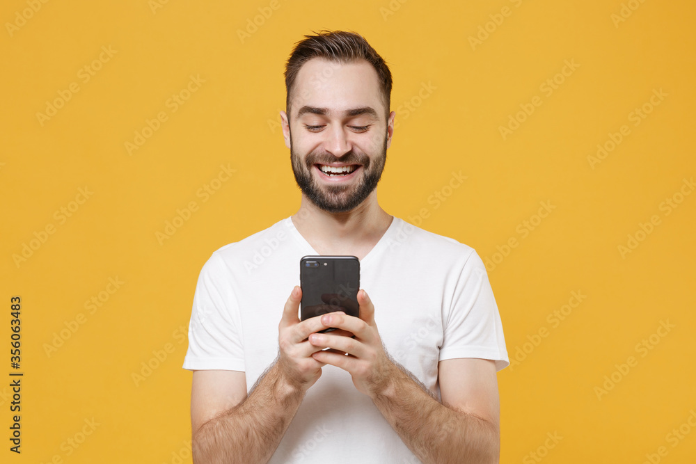 Smiling young bearded man guy in white casual t-shirt posing isolated on yellow wall background studio portrait. People lifestyle concept. Mock up copy space. Using mobile phone, typing sms message. Smiling young bearded man guy in white casual t-shirt posing isolated on yellow wall background studio portrait. People lifestyle concept. Mock up copy space. Using mobile phone, typing sms message.
