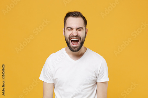 Crazy young bearded man guy in white casual t-shirt posing isolated on yellow background studio portrait. People sincere emotions lifestyle concept. Mock up copy space. Keeping eyes closed, screaming.