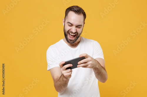 Cheerful young bearded man guy in white casual t-shirt posing isolated on yellow wall background studio portrait. People emotions lifestyle concept. Mock up copy space. Play game with mobile phone.