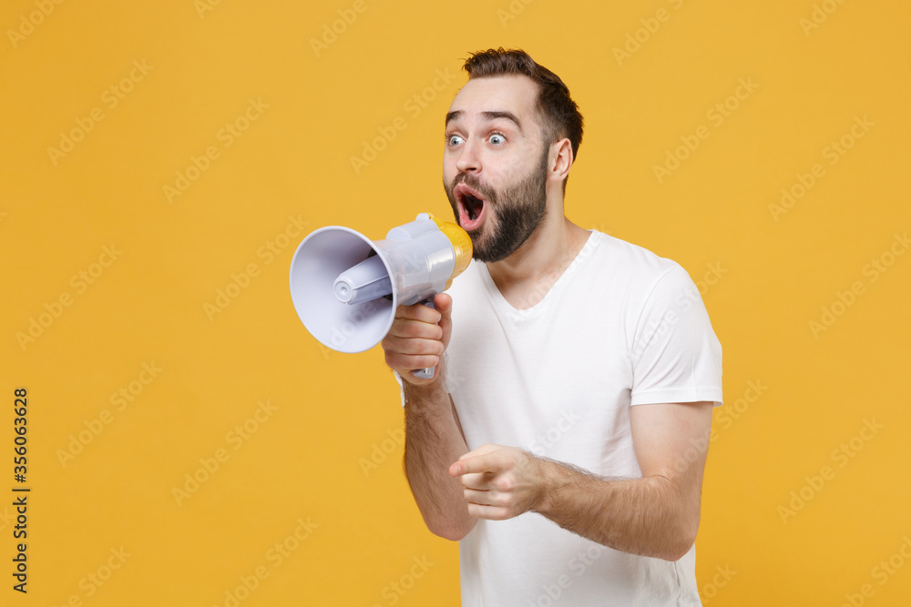 Shocked young bearded man guy in white casual t-shirt posing isolated on yellow background studio portrait. People lifestyle concept. Mock up copy space. Scream in megaphone point index finger aside. Shocked young bearded man guy in white casual t-shirt posing isolated on yellow background studio portrait. People lifestyle concept. Mock up copy space. Scream in megaphone point index finger aside.