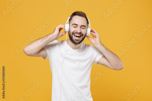 Cheerful young bearded man guy in white casual t-shirt posing isolated on yellow wall background studio. People lifestyle concept. Mock up copy space. Listen music with headphones keeping eyes closed.