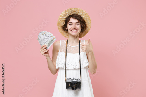 Smiling tourist girl in dress hat photo camera isolated on pink background. Traveling abroad weekend getaway. Air flight journey concept. Hold fan of cash money in dollar banknotes showing thumb up.