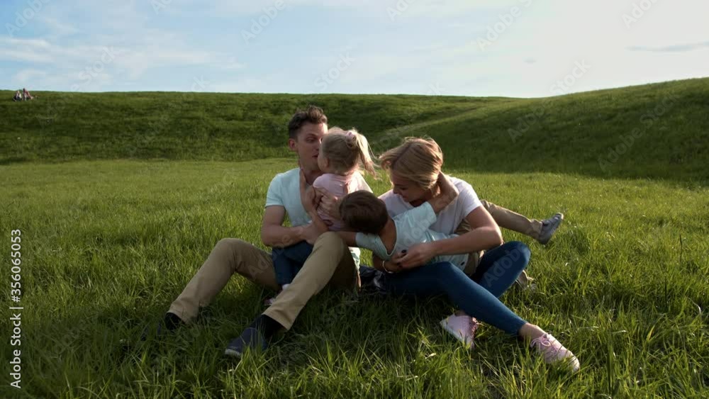 Young parents in clothes in pastel colors are sitting on the grass in the park, from different sides son and daughter come running to them, mom and dad hug them, and then they hug together Young parents in clothes in pastel colors are sitting on the grass in the park, from different sides son and daughter come running to them, mom and dad hug them, and then they hug together