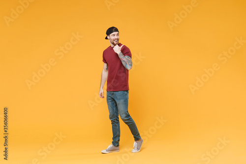 Cheerful young tattooed man guy in casual t-shirt black cap posing isolated on yellow background studio portrait. People sincere emotions lifestyle concept. Mock up copy space. Pointing thumb aside.