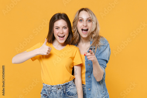 Two excited young women girls friends in casual t-shirts denim clothes isolated on yellow background. People lifestyle concept. Mock up copy space. Showing thumb up, pointing index finger on camera.