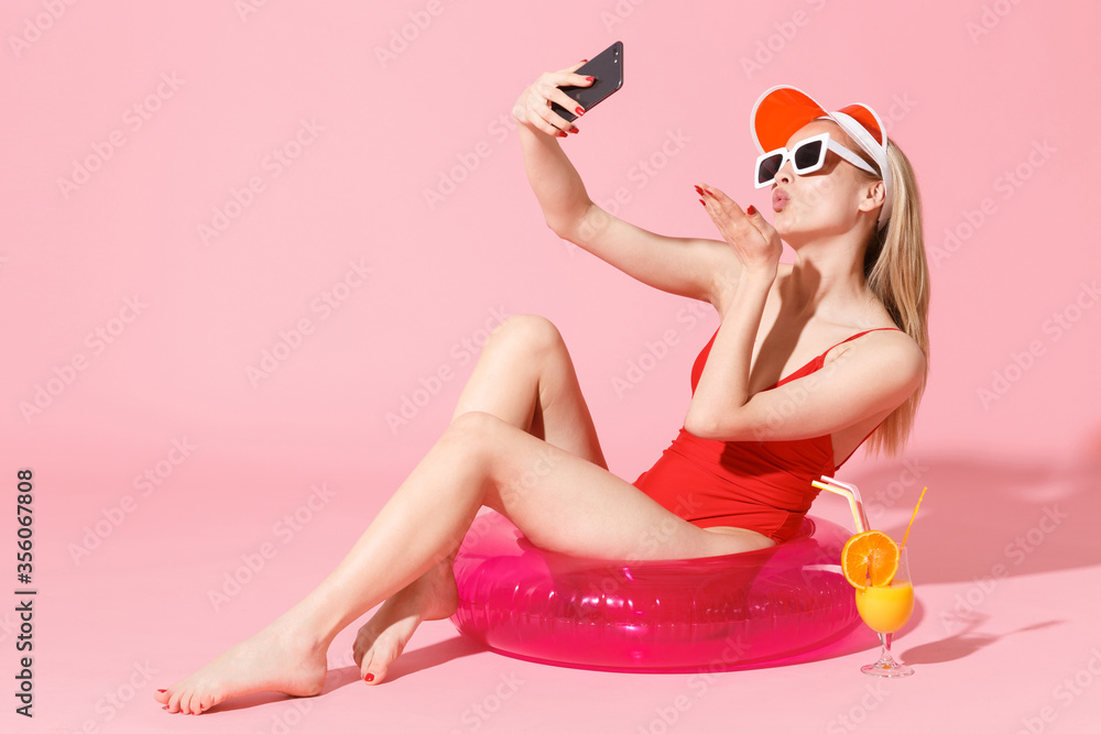 Charming woman girl in red one-piece swimsuit cap glasses isolated on pink background. People summer vacation rest lifestyle concept. Sit in swim inflatable ring doing selfie shot on mobile phone.