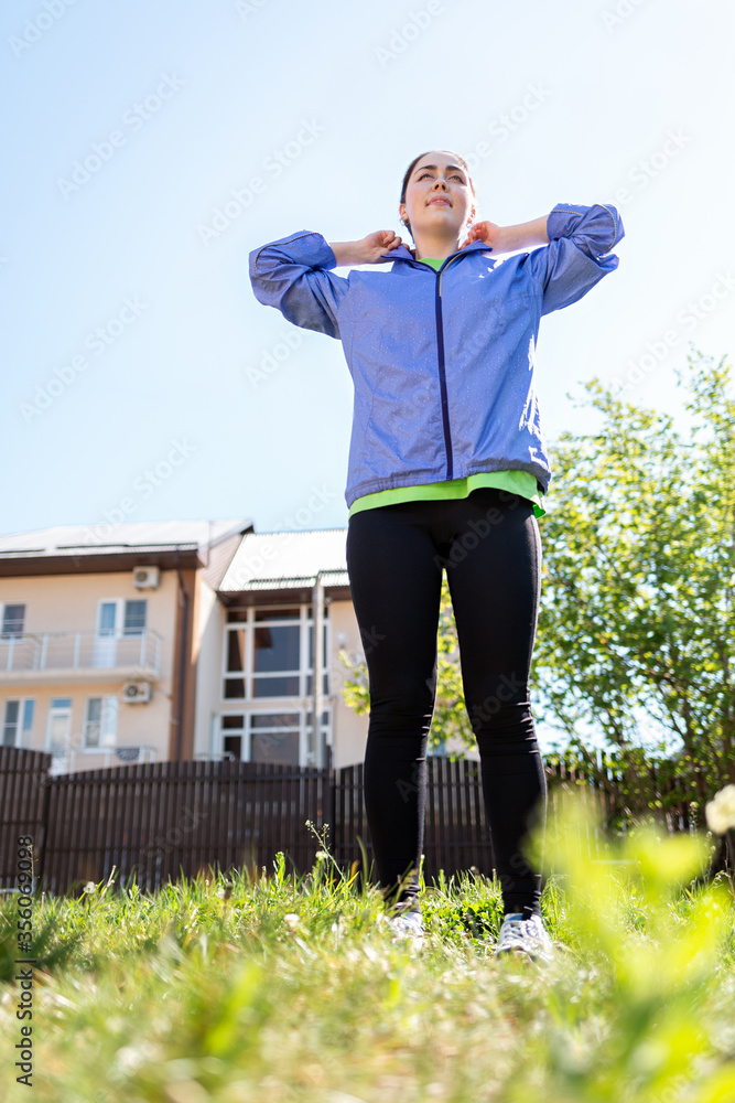 A happy young woman is doing sports in the yard, stretching her arms. View from below, from the grass. Concept of outdoor sports activity