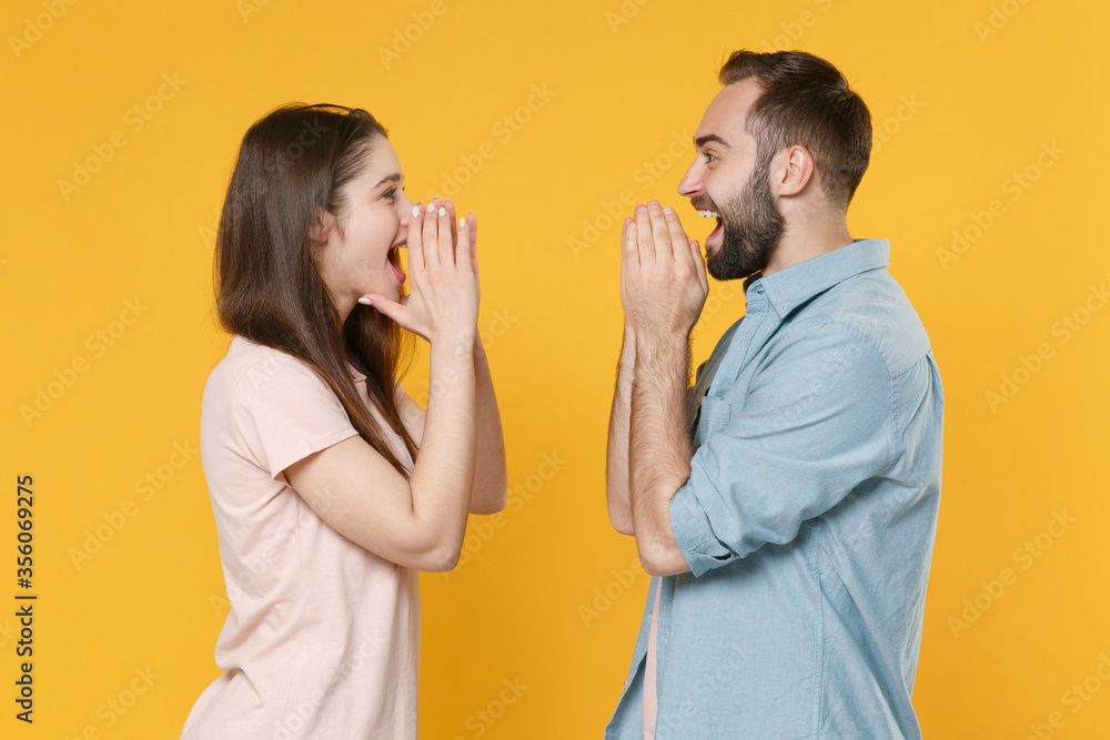 Side view excited young couple two friends guy girl in pastel blue casual clothes isolated on yellow background. People lifestyle concept. Scream with hands gesture near mouth, looking at each other. Side view excited young couple two friends guy girl in pastel blue casual clothes isolated on yellow background. People lifestyle concept. Scream with hands gesture near mouth, looking at each other.