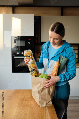 Portrait of young woman in sport werar holding craft paper bag with organic veggies, pasta and bred in at home in the kitchen photo