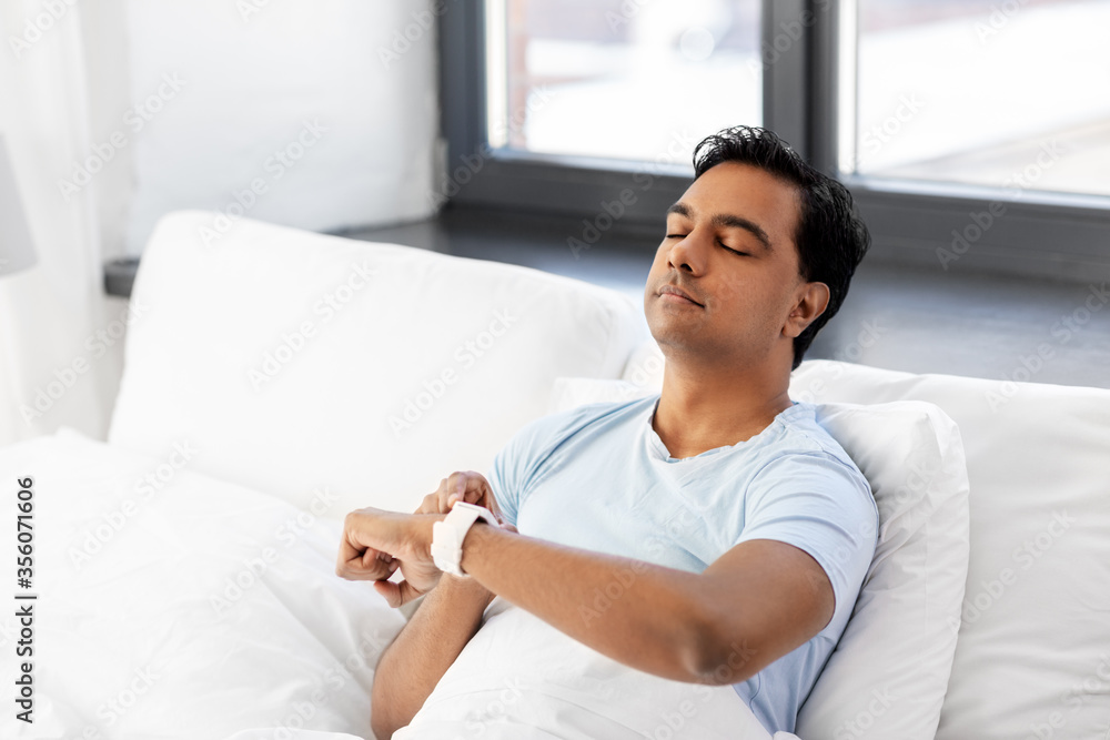 people, bedtime and rest concept - happy smiling indian man with smart watch doing breath exercise lying in bed at home people, bedtime and rest concept - happy smiling indian man with smart watch doing breath exercise lying in bed at home