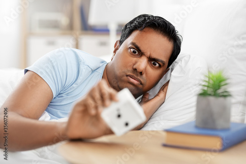 people, bedtime and rest concept - overslept indian man looking at alarm clock lying in bed at home photo