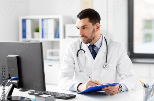 healthcare, medicine and people concept - male doctor with clipboard and computer working at hospital