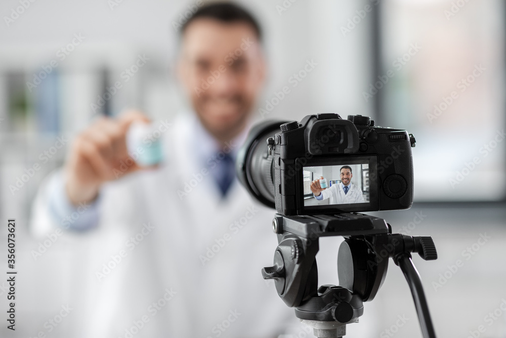 healthcare, medicine and blogging concept - happy smiling male doctor with camera showing drug and recording video blog at hospital