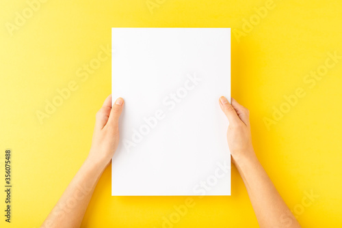 Young woman’s hands showing blank paper sheet on yellow desktop. Top view