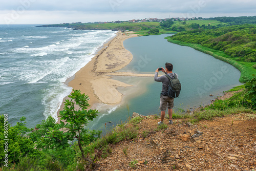 Fényképezés Rear view of a man taking pictures on his smart phone while standing at the edge of a rock looking toward beautiful and wild beach at the Black Sea coast and the estuary of Veleka river, Bulgaria
