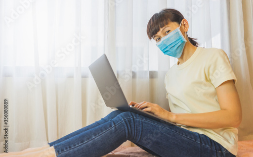Young woman wear a mask using laptop on bed at home. Work from home concept