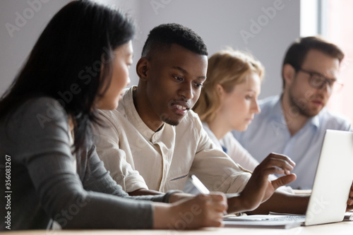 African and asian different age colleagues seated at desk in co-working room look at laptop screen discuss new corporate app, more experienced mate explain program software to intern mentoring concept