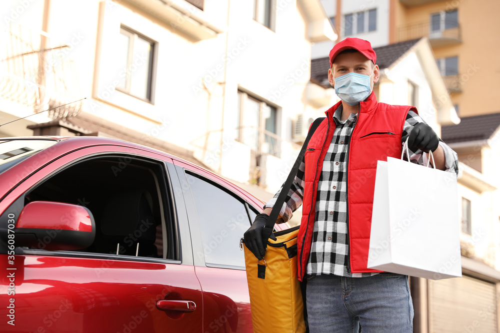 Courier in protective mask with order and thermobag near car outdoors. Food delivery service during coronavirus quarantine Courier in protective mask with order and thermobag near car outdoors. Food delivery service during coronavirus quarantine