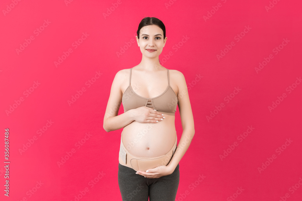 Portrait of pregnant woman using pregnancy belt at pink background with copy space. Orthopedic abdominal support belt concept Portrait of pregnant woman using pregnancy belt at pink background with copy space. Orthopedic abdominal support belt concept