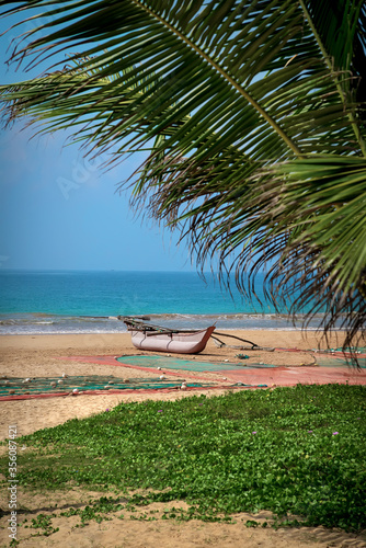 Fototapeta Naklejka Na Ścianę i Meble -  Fisher wooden boat lying on saecoast, palm leaf is in frame. Fishnet is laying on a sand. Relaxing scenery of asian tropical island. Exotic travel, summertime holidays.