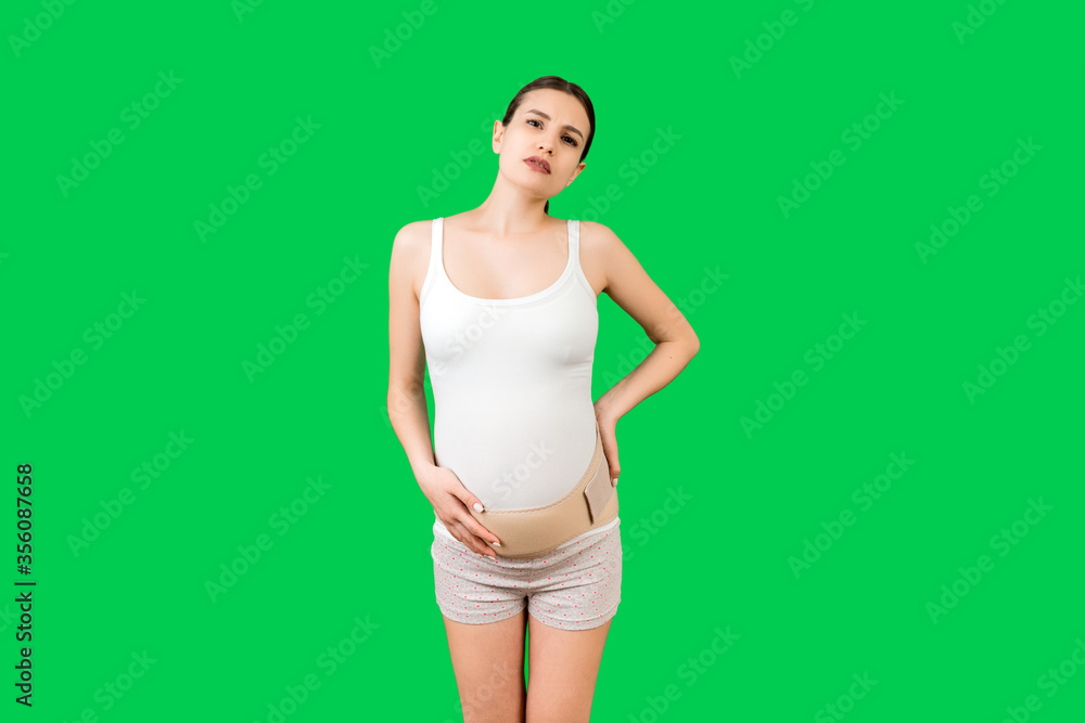 Portrait of supporting bandage against backache on pregnant woman at green background with copy space. Mother is suffering from pain in the back. Orthopedic abdominal support belt concept Portrait of supporting bandage against backache on pregnant woman at green background with copy space. Mother is suffering from pain in the back. Orthopedic abdominal support belt concept