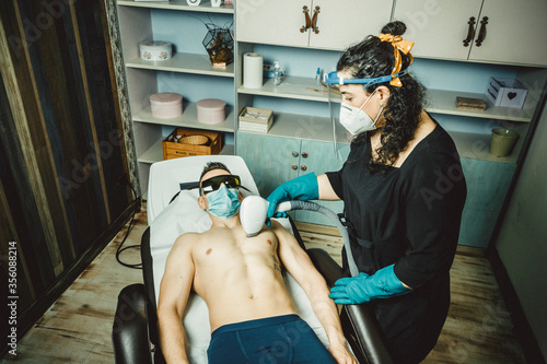 Mujer realizando depilación laser a hombre en centro de belleza y estética con las medidas de protección personal puestas, mascarilla y pantalla, por el virus COVID-19 photo