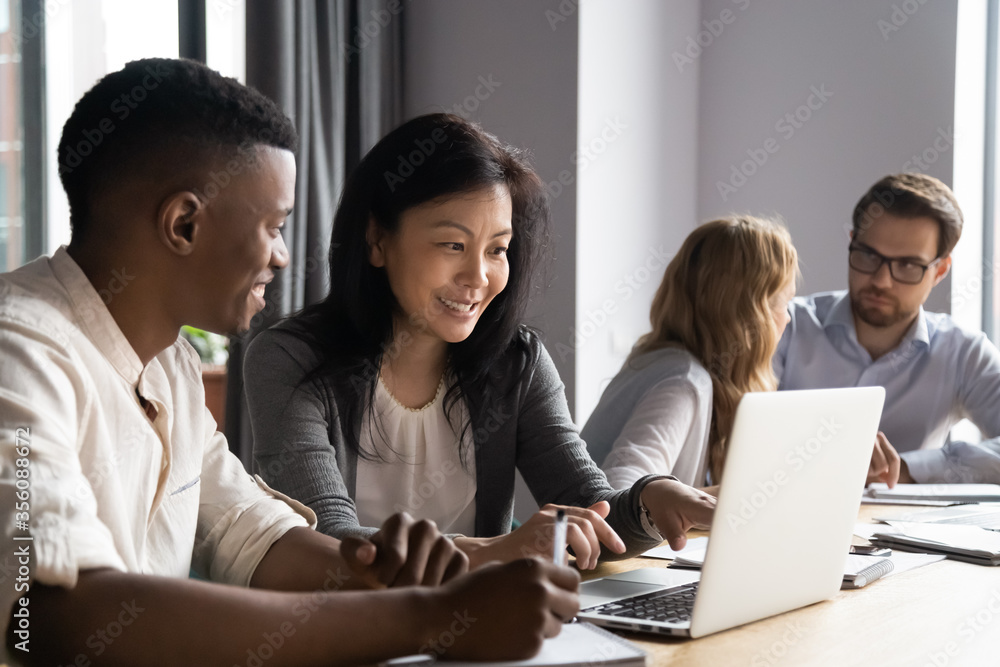 Multi-ethnic affiliates working on task together seated at desk in co-working, mature asian mentor teach new employee african guy, diverse mates having fun using laptop, learn corporate apps concept Multi-ethnic affiliates working on task together seated at desk in co-working, mature asian mentor teach new employee african guy, diverse mates having fun using laptop, learn corporate apps concept