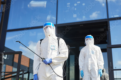 Low angle view at two workers wearing protective suits posing with disinfection gear outdoors while standing against glass building, copy space photo