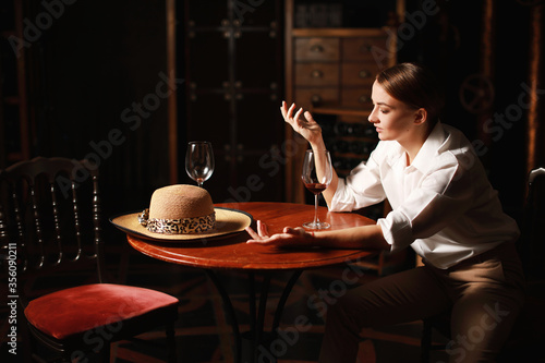 Beautiful theatrical actress woman with retro fashion style on the stage siting near wooden table with glass of red wine and vintage hat. 
