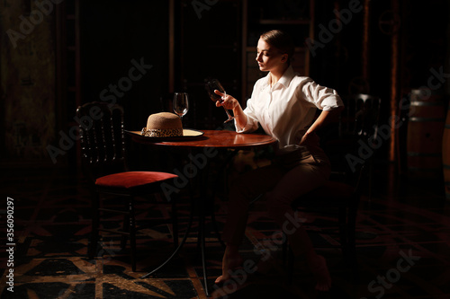 Beautiful theatrical actress woman with retro fashion style on the stage siting near wooden table with glass of red wine and vintage hat. 