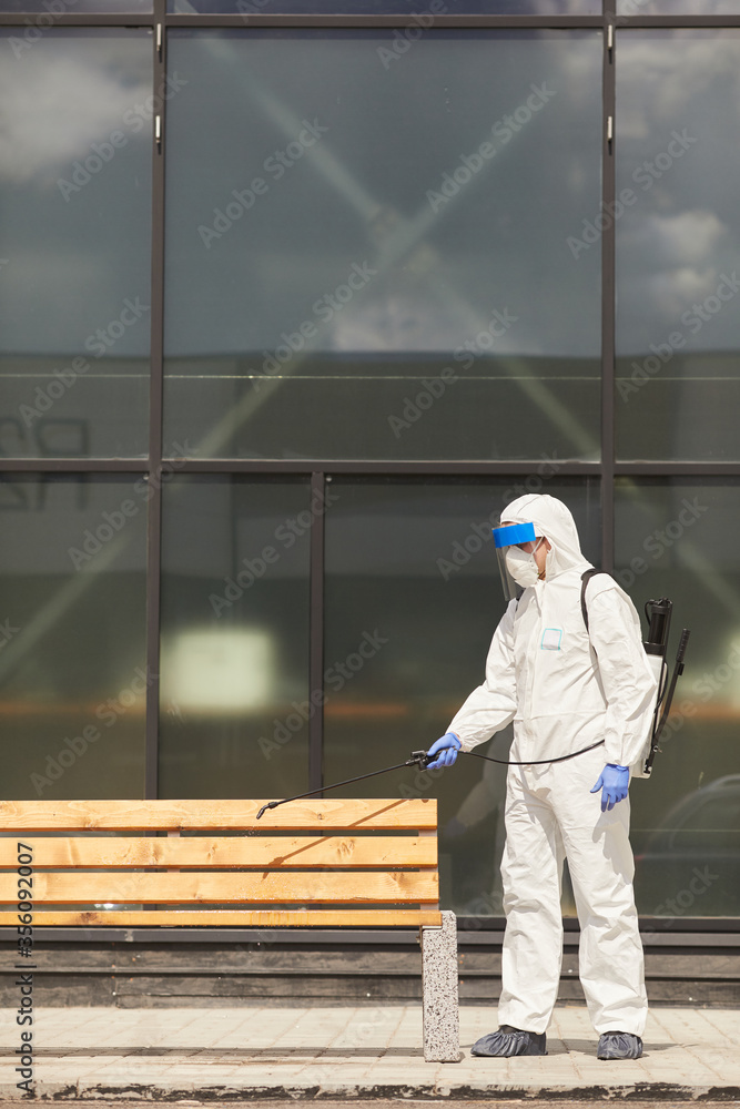 Side view full length portrait of one worker wearing protective suit spraying chemicals over bench outdoors during disinfection or cleaning, copy space Side view full length portrait of one worker wearing protective suit spraying chemicals over bench outdoors during disinfection or cleaning, copy space