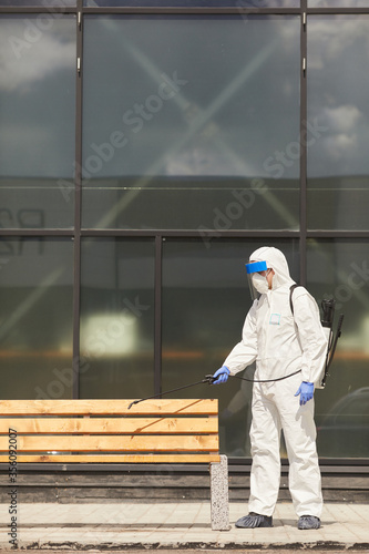 Side view full length portrait of one worker wearing protective suit spraying chemicals over bench outdoors during disinfection or cleaning, copy space