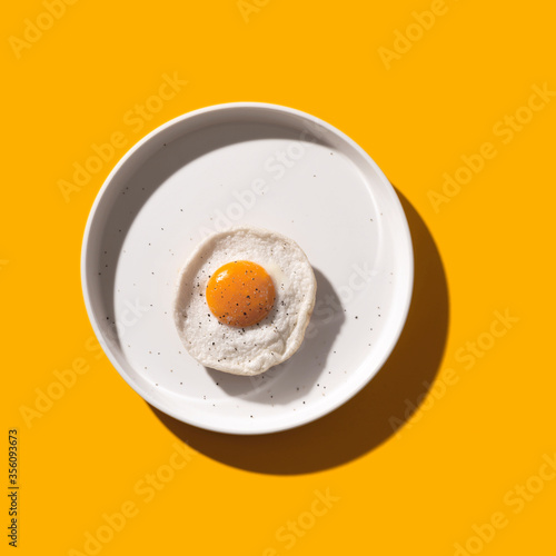 Fried egg on a round plate on a yellow table with hard shadows. The concept of creativity and minimalism in cooking. Top view, flat lay.