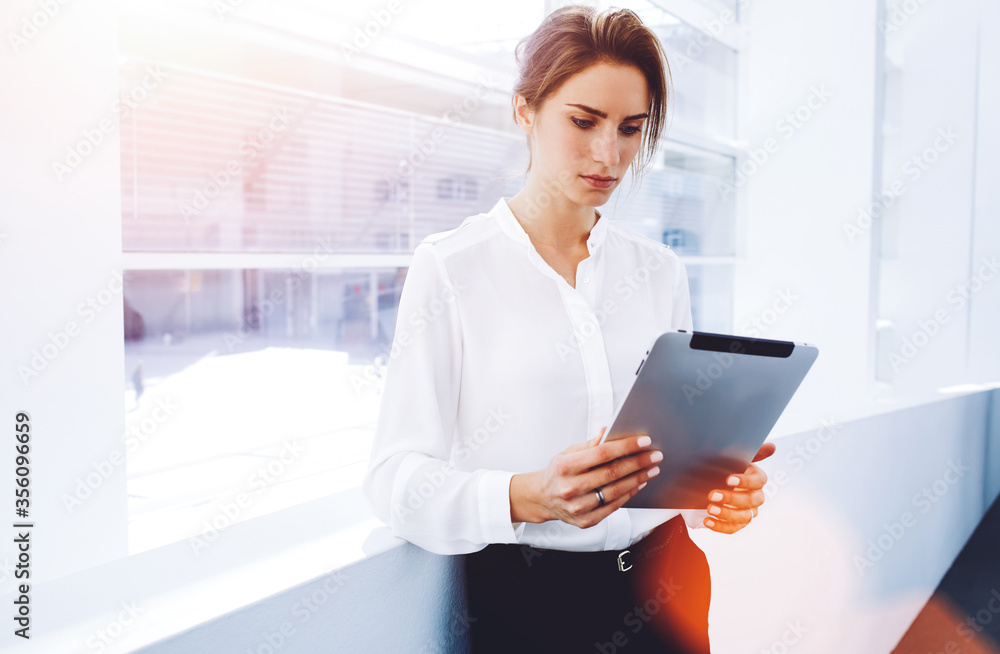 © BullRun - Young serious businesswoman using touch pad while standing in office interior near window