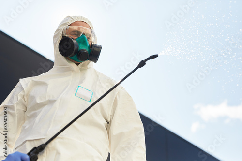 Low angle portrait of male worker wearing hazmat suit holding disinfection gear spraying chemicals outdoors while standing against sky background, copy space