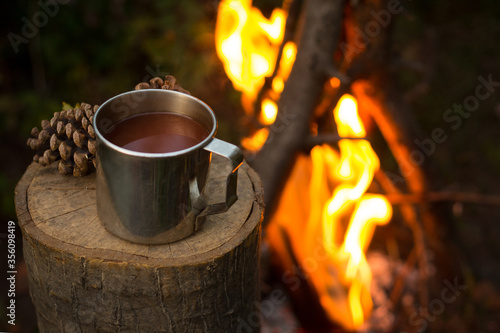 Travel tin cup with cocoa, chocolate, coffee on an old log by an outdoor campfire on background with pine cones. Lunch during the journey in the forest. Autumn mood. With extremely soft focus