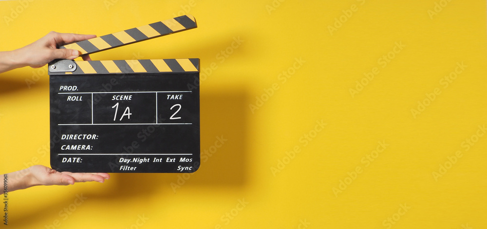 A Hand is holding Black clapperboard or movie slate on yellow background.It has written a number..