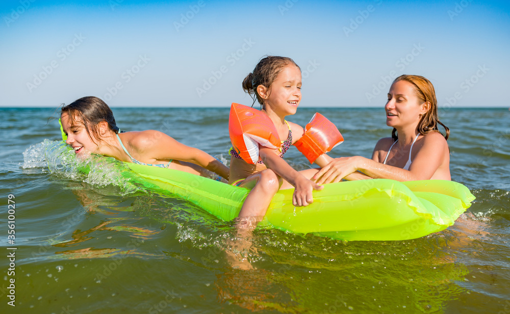 Obraz premium Happy mother and two small positive daughters bathe and swim in the sea with an air mattress on a sunny summer day. Vacation concept with kids. Copyspace